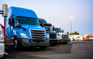 Different makes and models industrial grade professional big rigs semi trucks standing in row on the truck stop parking lot at evening time waiting to continued delivery schedule
